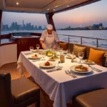 A chef in white uniform arranging gourmet dishes on a elegantly set dining table aboard a private yacht rental in Dubai, overlooking the city skyline at sunset.